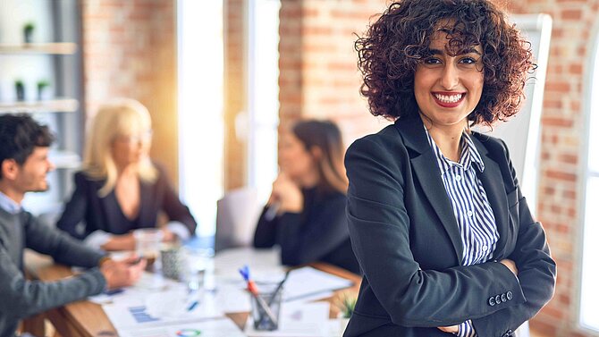 A woman in a business look stands in front of some colleagues in the office