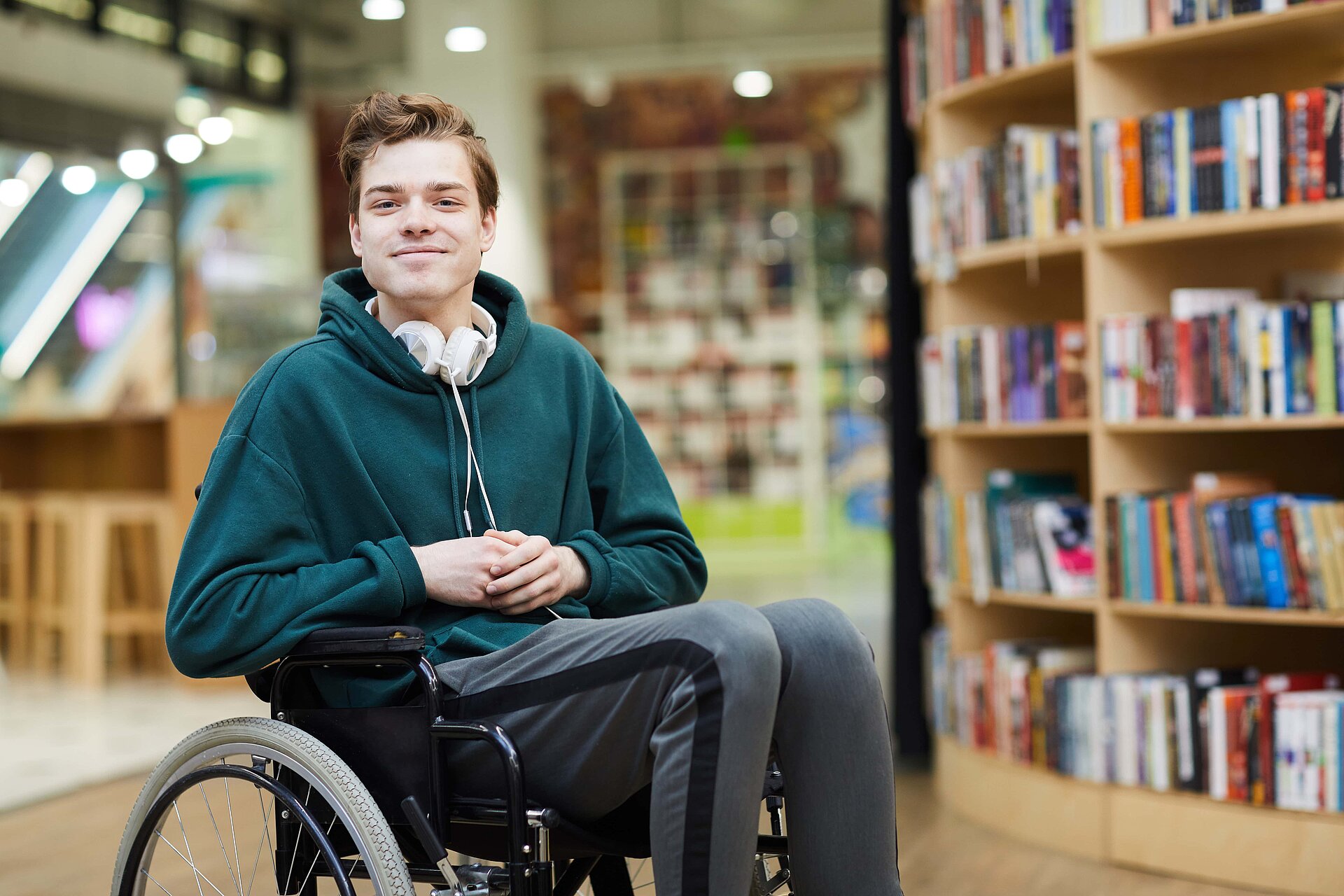 Spokojený, pohledný mladý student na vozíku se sluchátky na krku, který sedí v moderní knihovně nebo knihkupectví a dívá se do kamery.